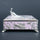 A decorative rectangular box with a lavender-pink enamel-like finish and raised silver-tone peacock and floral motifs, topped with a sculpted peacock figure on a dark gray background.