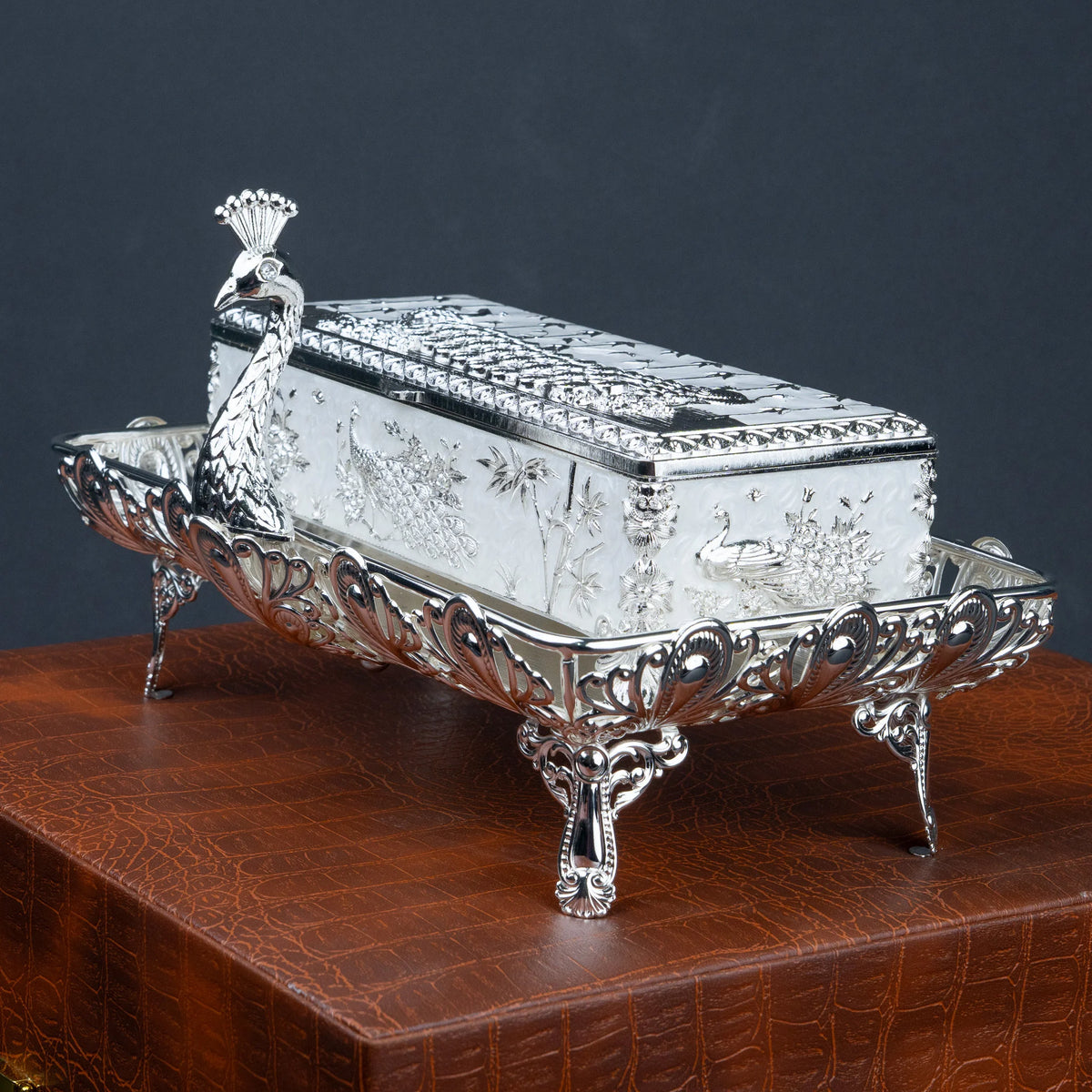 An ornate silver-toned decorative box with a lid, shown on a carved wooden pedestal against a dark gray background. The box features floral engravings and a peacock figure on the front.