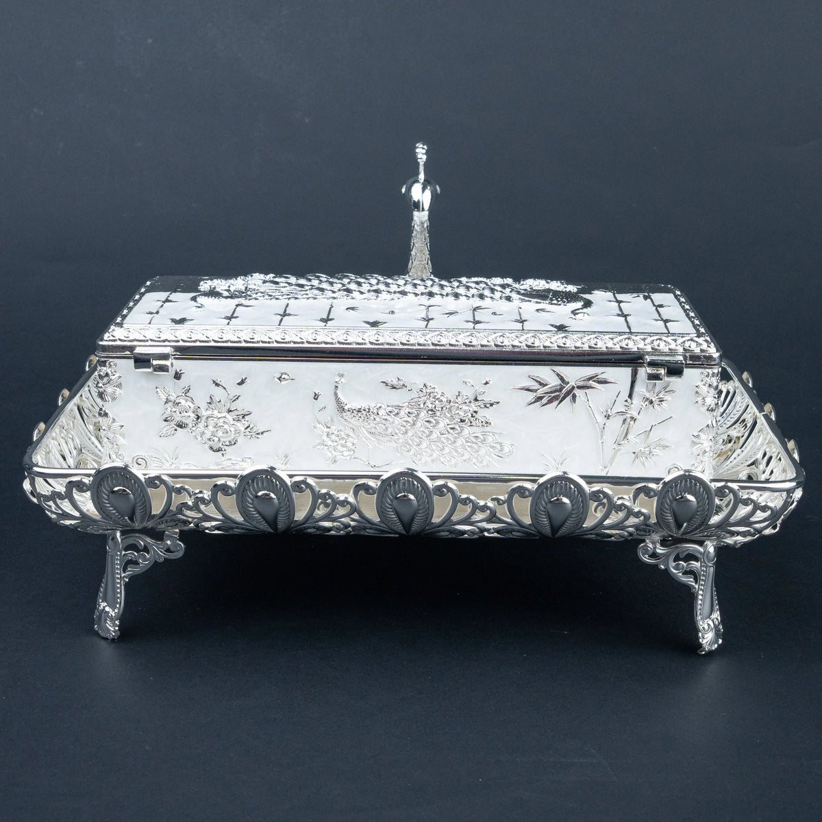 An ornate silver rectangular lidded box with decorative floral and bird engravings, raised feet, and a small standing figure on top, photographed against a dark studio background.