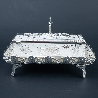An ornate silver rectangular lidded box with decorative floral and bird engravings, raised feet, and a small standing figure on top, photographed against a dark studio background.