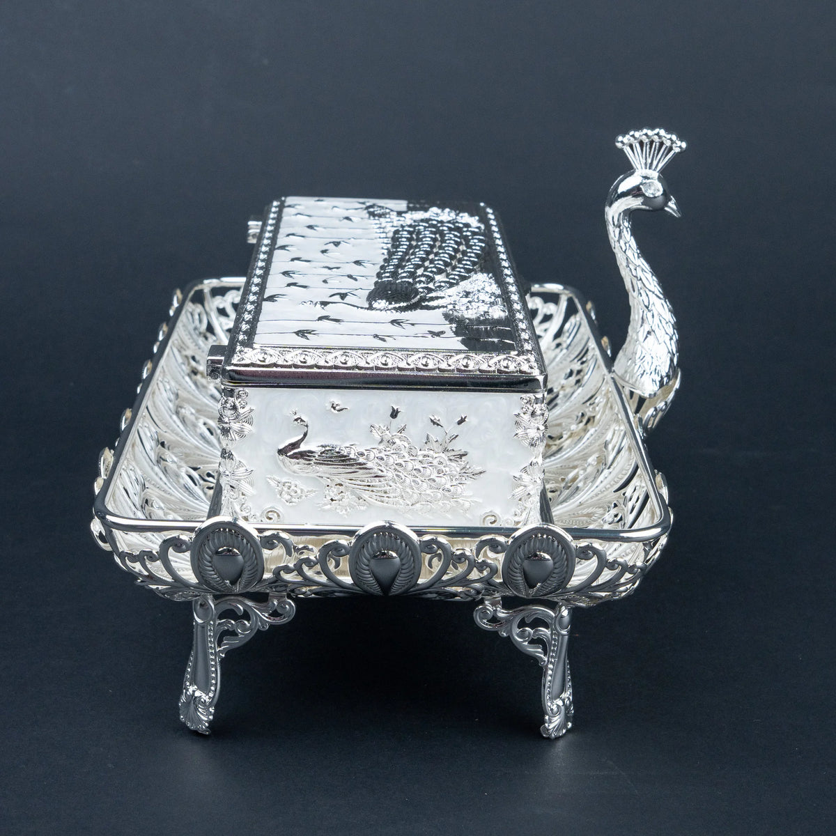 An ornate sterling silver peacock-shaped serving tray with a matching lid, featuring detailed engravings of peacocks and birds, set against a dark background.