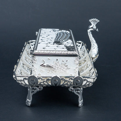 An ornate sterling silver peacock-shaped serving tray with a matching lid, featuring detailed engravings of peacocks and birds, set against a dark background.