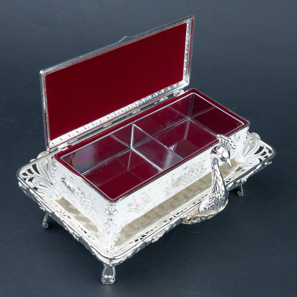 An ornate silver-toned jewelry box with a hinged lid, shown open to reveal a deep red velvet-lined interior divided into multiple compartments. The exterior features intricate floral engraving and a decorative handle, set against a dark gray background.