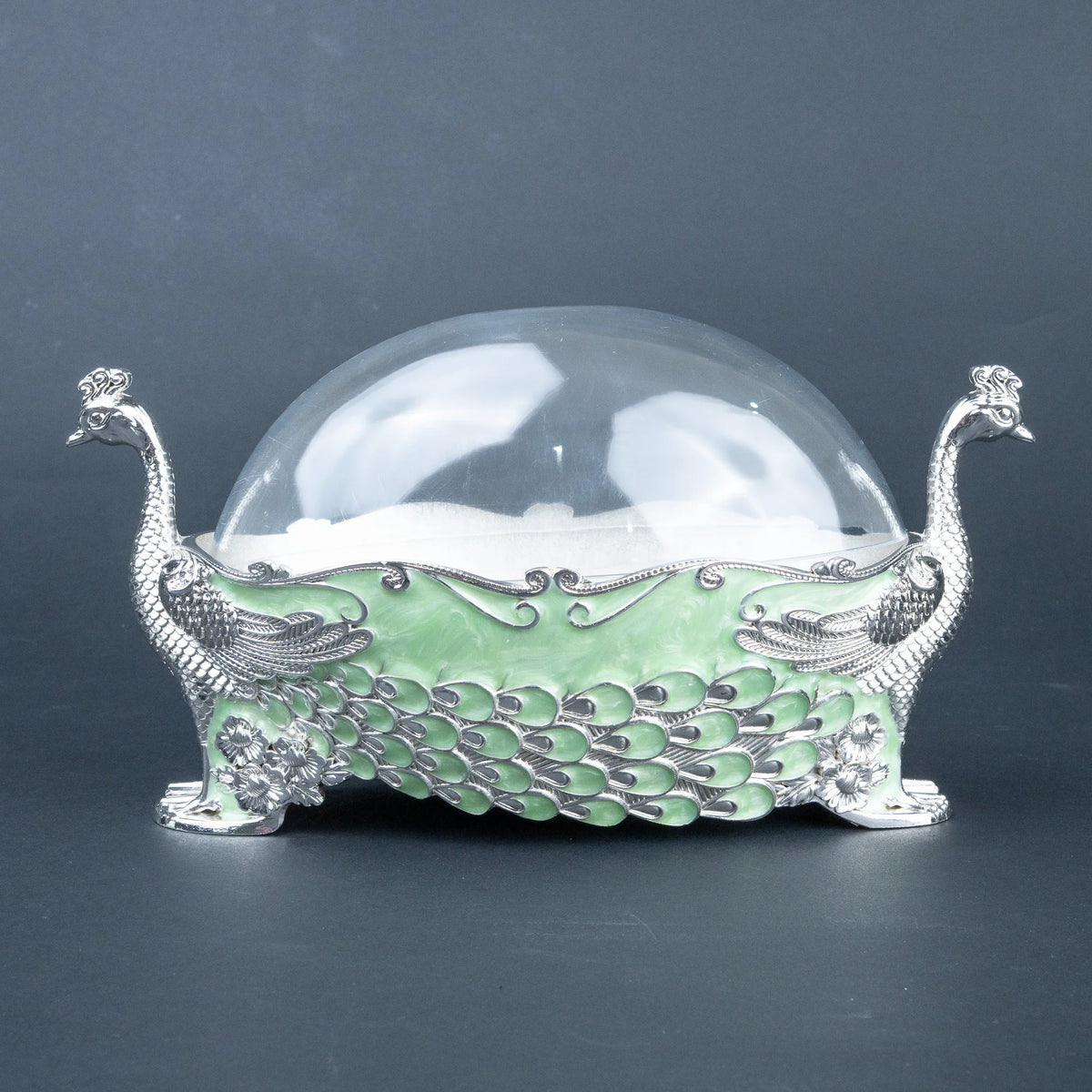 A decorative peacock-shaped container with a clear glass dome lid, featuring a light green body with silver-toned peacock feather detailing and two peacock heads at the sides, set against a dark gray background.