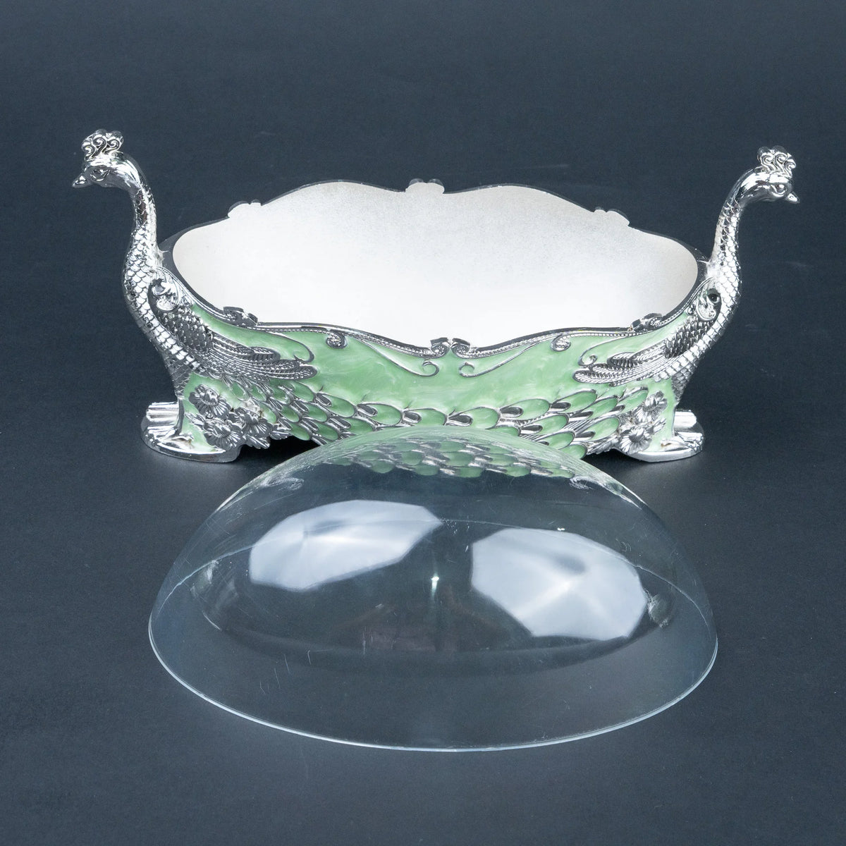 An ornate silver-toned peacock-shaped dish with a light green enamel body and a clear dome lid placed in front, photographed on a dark gray background.