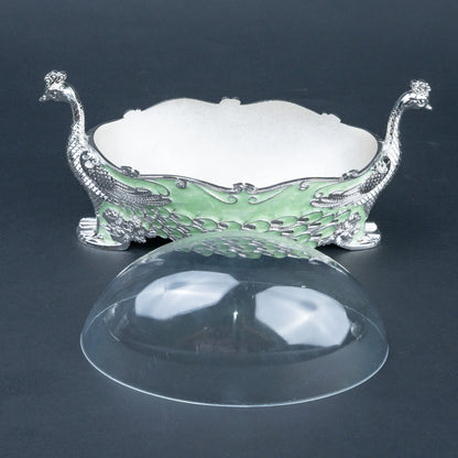 An ornate silver-toned peacock-shaped dish with a light green enamel body and a clear dome lid placed in front, photographed on a dark gray background.