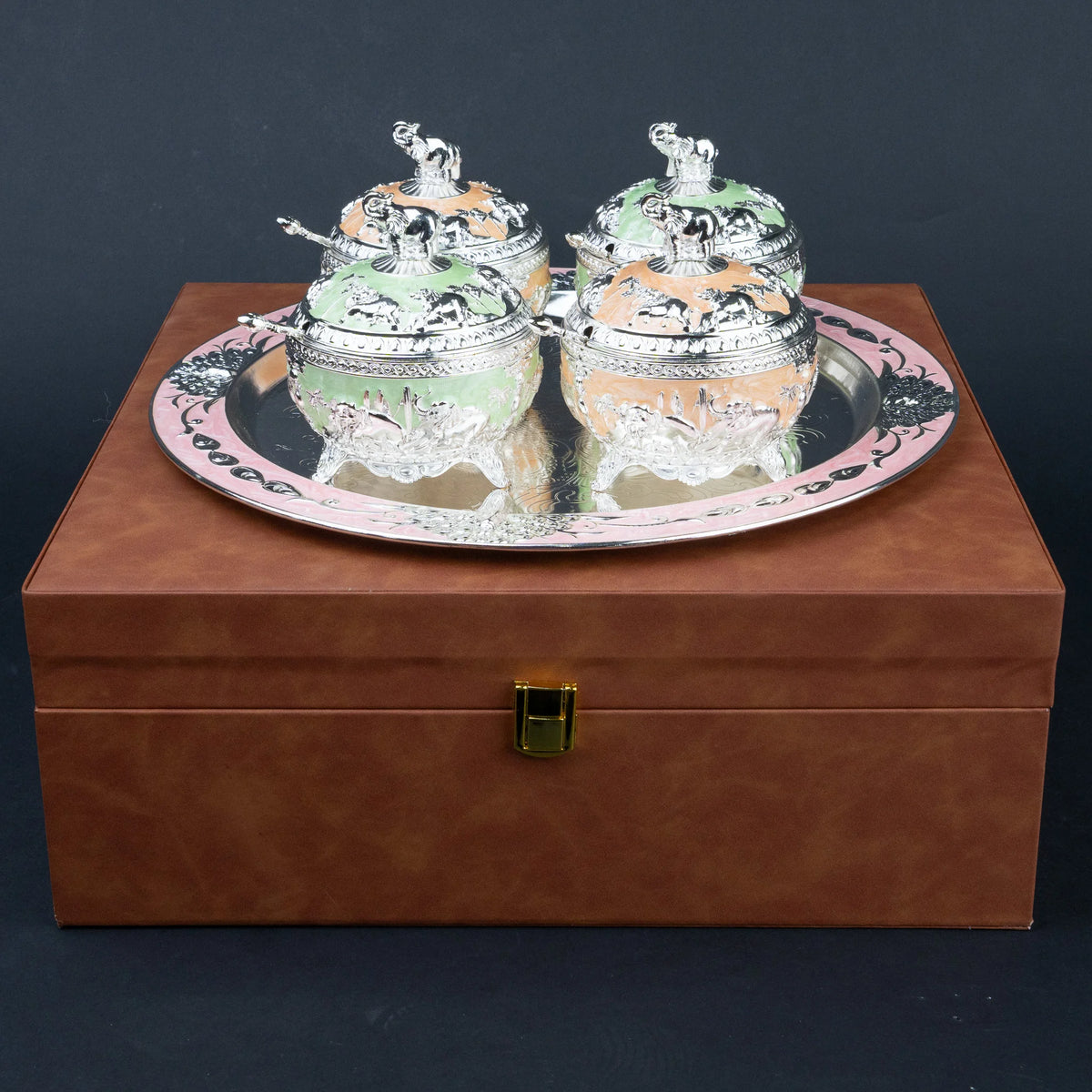 A pair of ornate silver-toned decorative boxes with colorful pastel inserts, displayed on a round silver tray atop a brown presentation box.