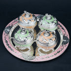 Four ornate lidded glass jars with silver-toned animal engravings and pastel orange and green interiors, arranged on a round pink and silver decorative tray against a black background.