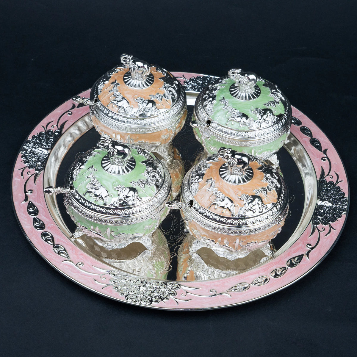 Four ornate lidded glass jars with silver-toned animal engravings and pastel orange and green interiors, arranged on a round pink and silver decorative tray against a black background.