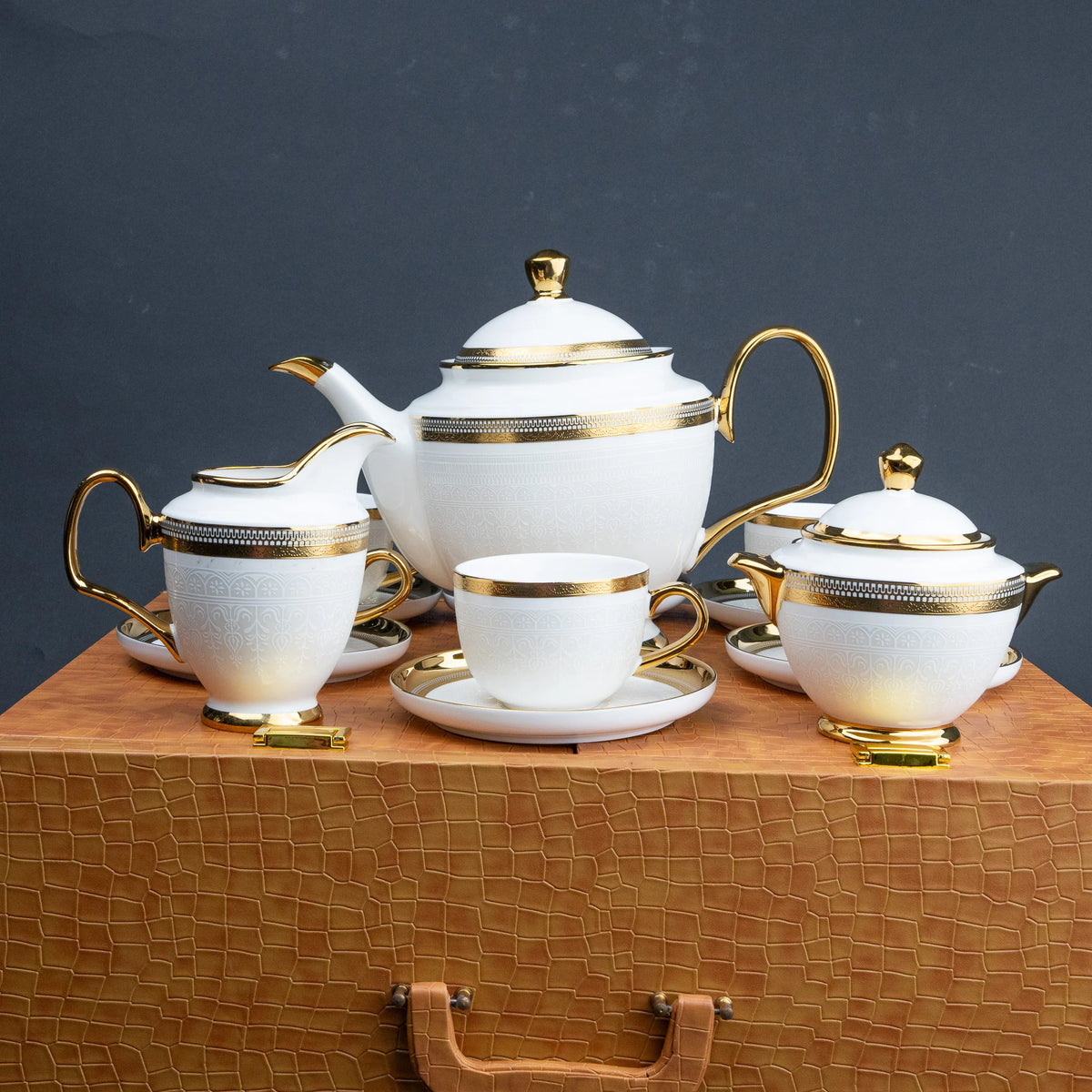 White porcelain tea set with gold trim, including a teapot, teacup, sugar bowl, and creamer, arranged on a textured brown surface against a dark gray background.