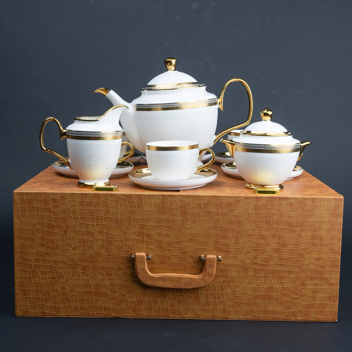 White ceramic tea set with gold trim, including a teapot, cups, saucers, and a sugar bowl, arranged on a textured brown gift box against a dark gray background.