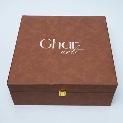 A square brown faux-leather gift box with a gold clasp and the brand name 'Ghar art' printed in white on the lid, set against a light gray background.