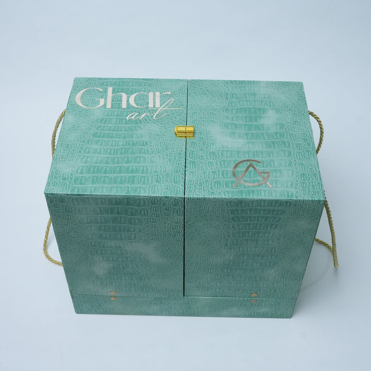 A square mint green gift box with a crocodile-embossed texture, featuring the brand name 'Ghar art' in white and gold on the top left, a small gold clasp in the center, and gold rope handles on the sides.
