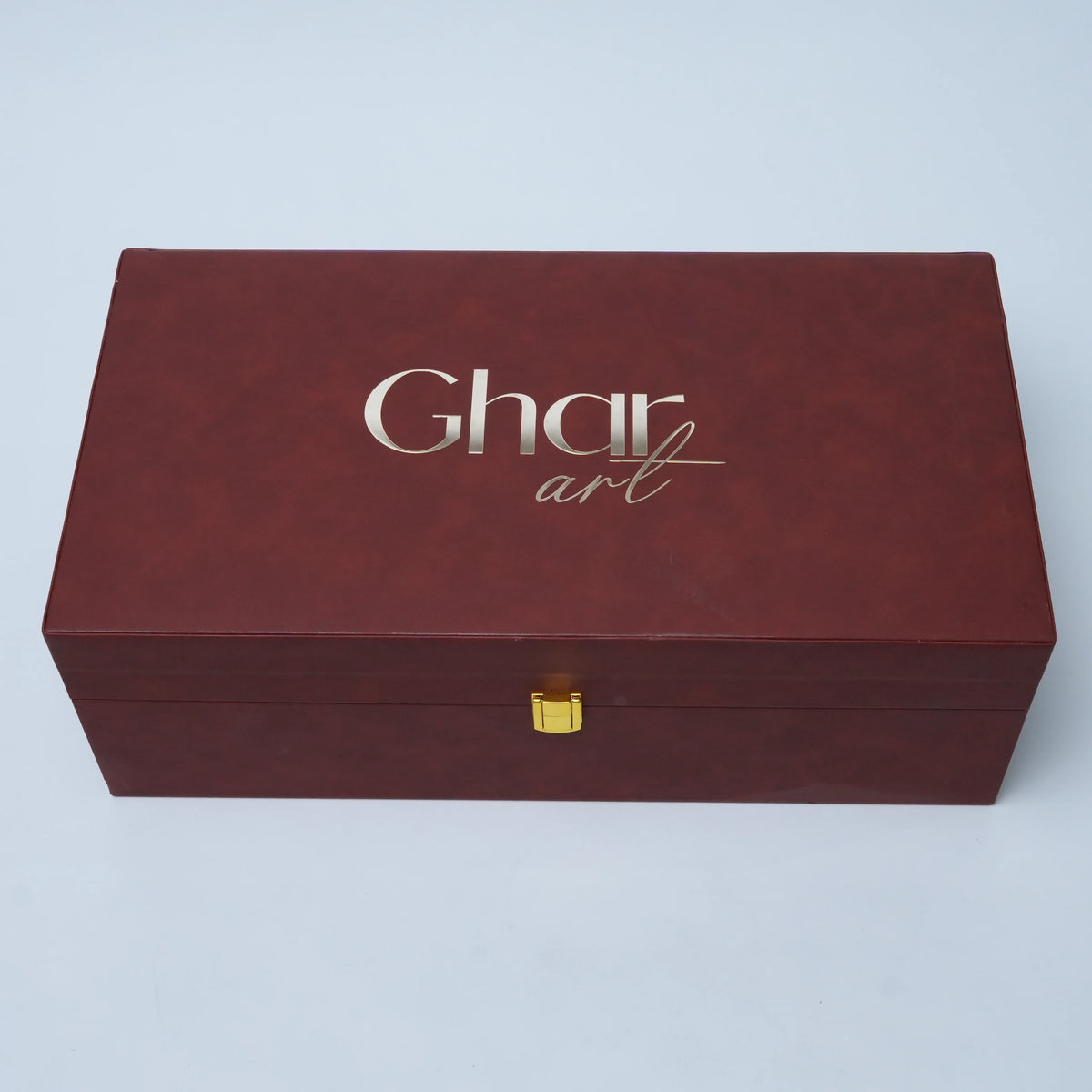A rectangular dark reddish-brown faux-leather gift box with a gold clasp and silver 'Ghar art' branding on the lid, set against a light gray background.