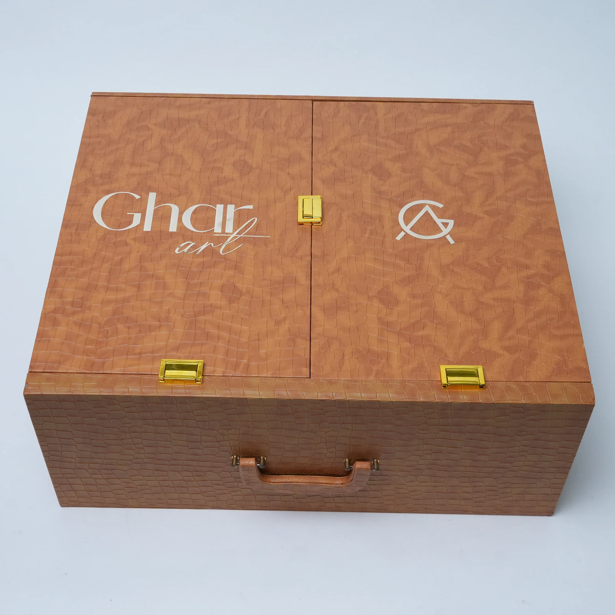 A rectangular wooden presentation box with two hinged doors, featuring a warm brown woodgrain finish, gold-tone metal latches, a small leather handle on the front, and white printed branding on the lid that reads 'Ghar art' alongside a stylized symbol.