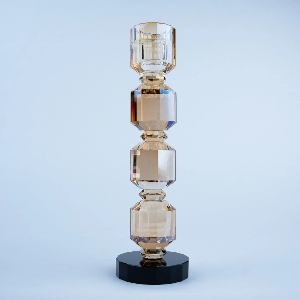 A modern geometric crystal candlestick holder with four stacked faceted glass sections in a warm champagne color, resting on a round black base against a light blue-gray studio background.