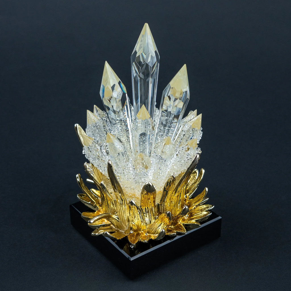 A decorative crystal cluster sculpture with clear faceted crystal points rising from a textured white center and a metallic gold lotus-like base, set on a black square pedestal against a dark background.