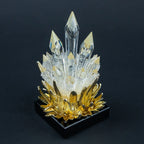 A decorative crystal cluster sculpture with clear faceted crystal points rising from a textured white center and a metallic gold lotus-like base, set on a black square pedestal against a dark background.