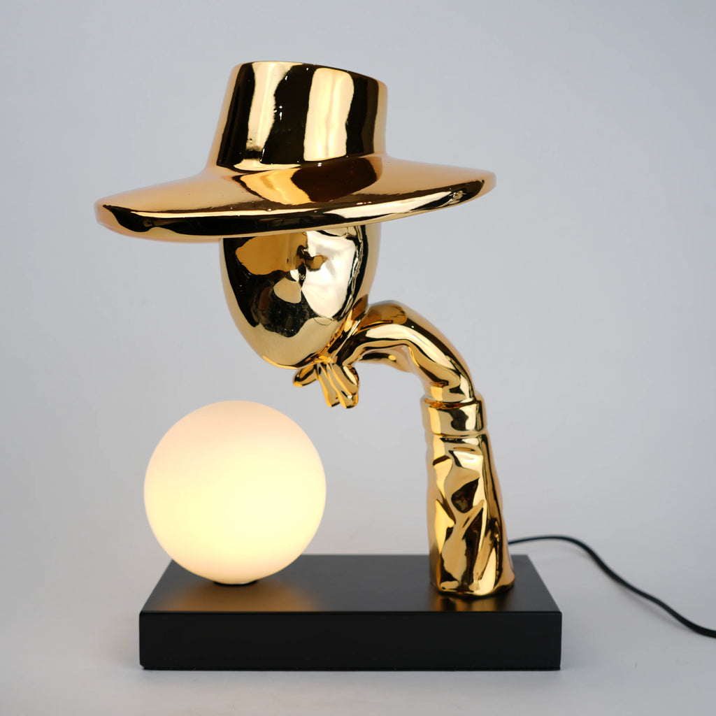 Gold electroplated table lamp featuring a sculptural curved human-like form with a wide hat-shaped top and a glowing spherical light on a rectangular base, designed with a smooth reflective metallic surface.