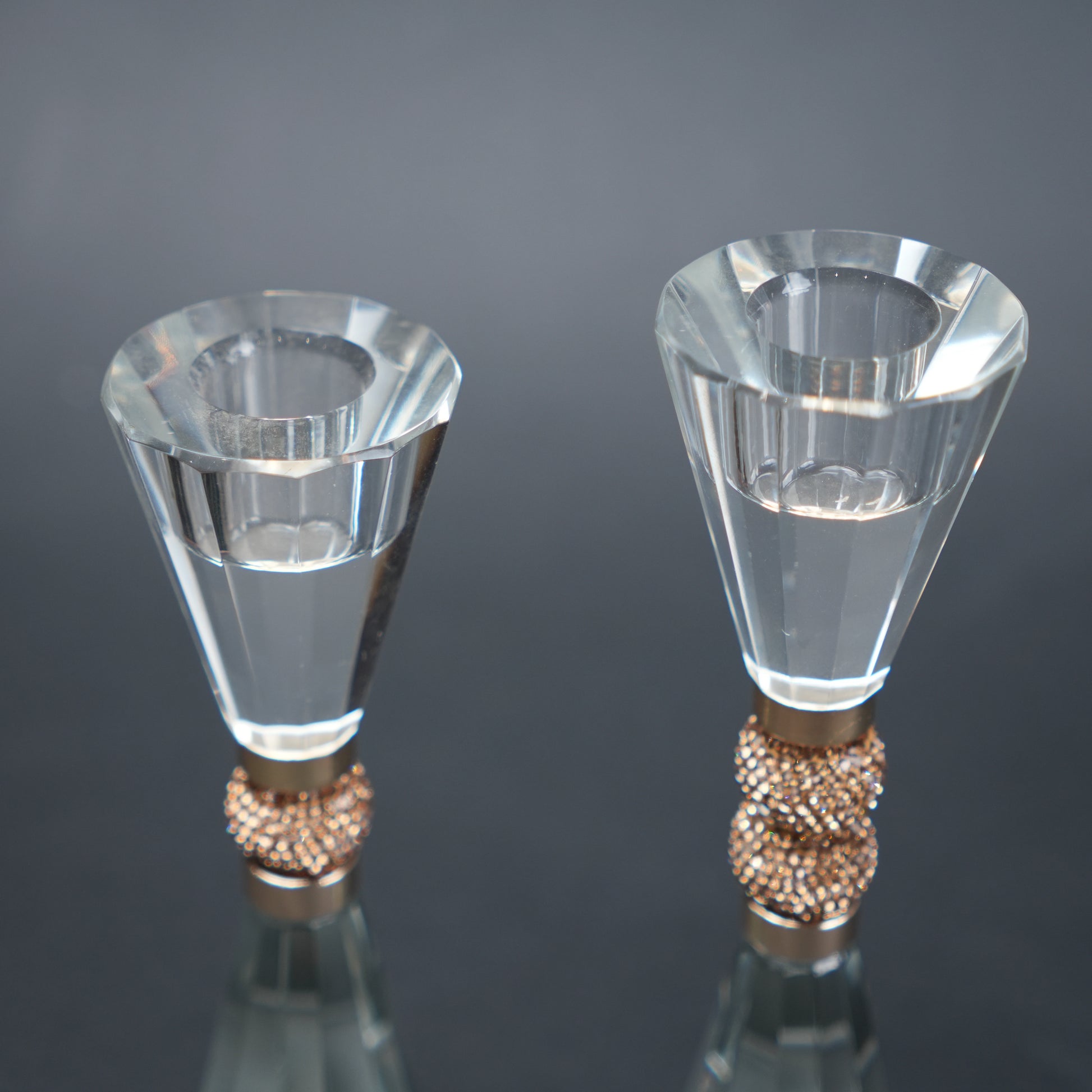 Two clear crystal candle holders with rose gold bases on a reflective surface.
