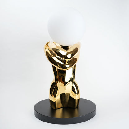 Gold abstract sculpture with a white sphere on a black base against a white background