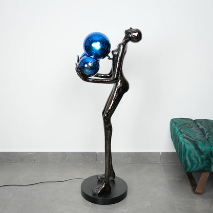 Modern sculpture of a figure holding blue spheres against a white wall.