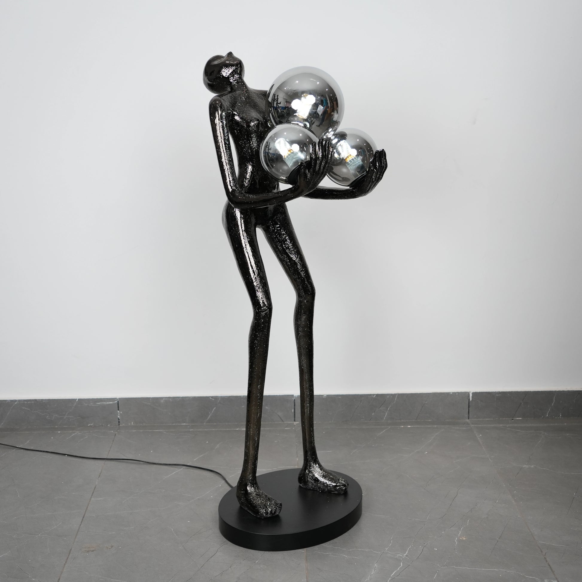 Black sculpture of a figure holding three reflective spheres on a gray floor.