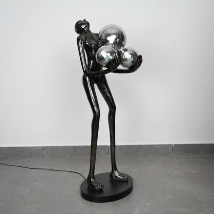 Black sculpture of a figure holding three reflective spheres on a gray floor.