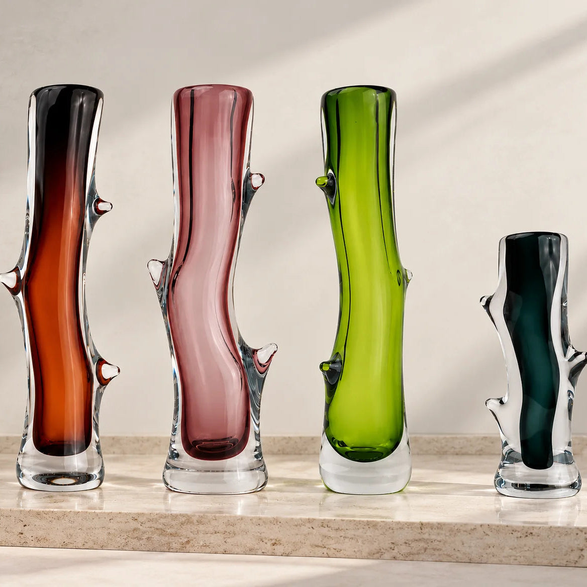 Set of four artistic sculptural glass vases in dark brown, blush pink, lime green, and charcoal, featuring glossy transparent glass bases, elegant curved silhouettes, and unique organic branch-like detailing, displayed together on a clean white studio background.