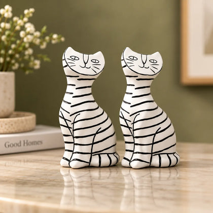 Two resin cat figurines with black stripes on a white glaze, displayed side-by-side on a polished marble countertop in a styled home setting.