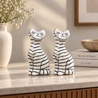 Two white resin cat figurines with black stripes sitting on a light marble countertop in a styled home interior.