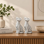 Two white resin cat figurines with black horizontal stripes sit on a wooden console table in a styled interior setting with a vase of green leaves and books.
