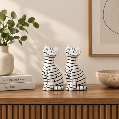 Two white resin cat figurines with black horizontal stripes sit on a wooden console table in a styled interior setting with a vase of green leaves and books.