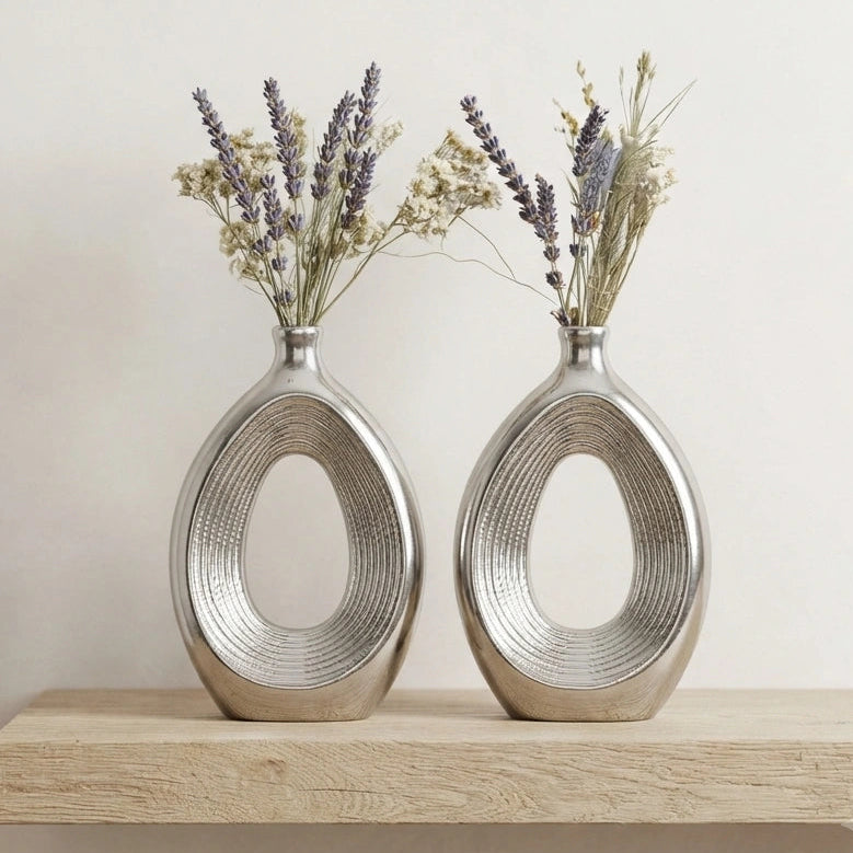 Two metallic silver oval vases with a textured ribbed design, filled with dried lavender and baby's breath, displayed on a light wood surface against a neutral beige wall.