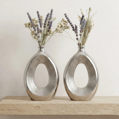Two metallic silver oval vases with a textured ribbed design, filled with dried lavender and baby's breath, displayed on a light wood surface against a neutral beige wall.