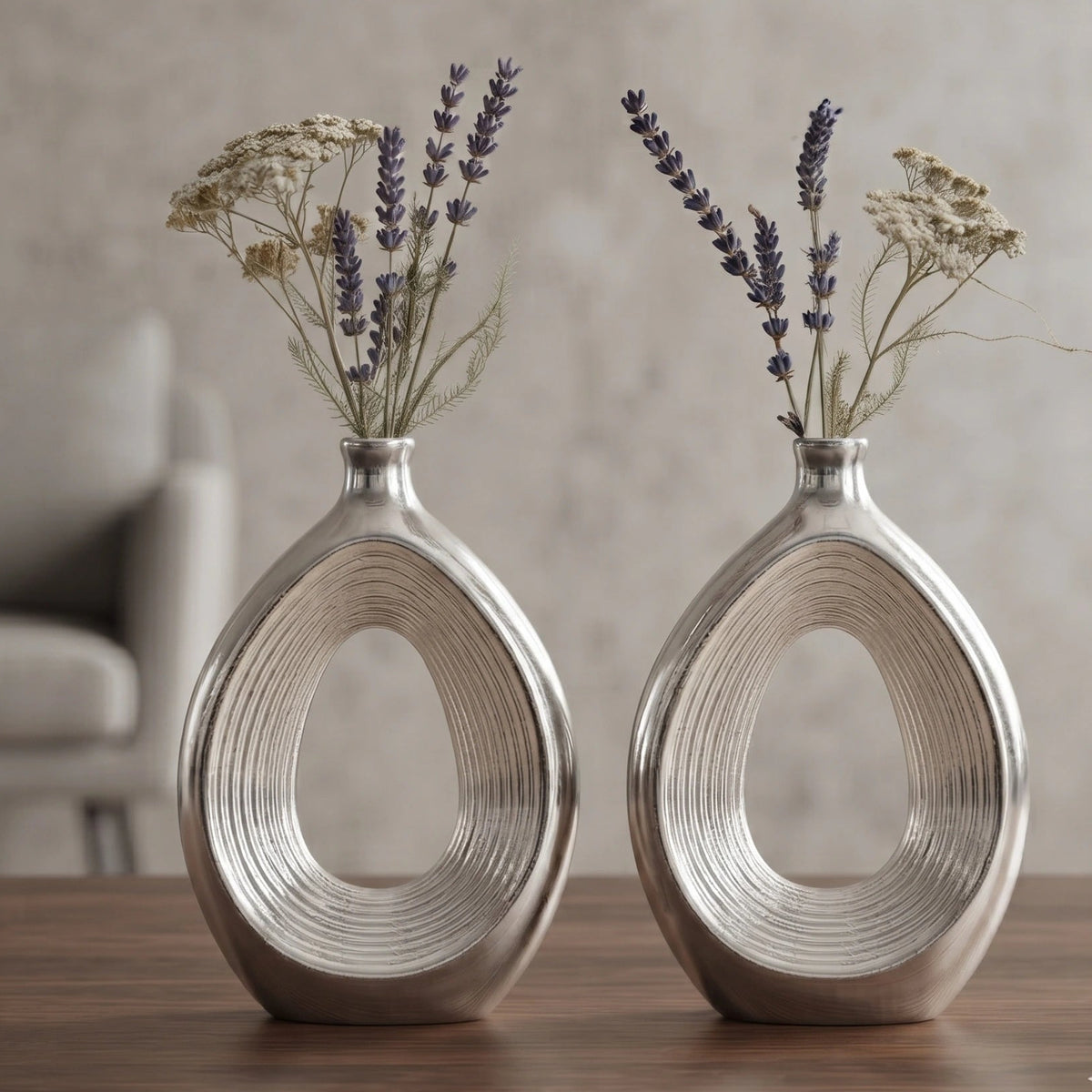 Two matching metallic silver oval vases with a ribbed textured finish, each filled with dried lavender and white flowers, displayed on a wooden surface against a neutral beige background.