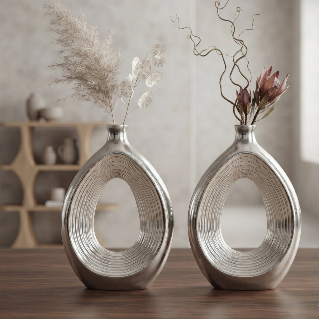 Two matching metallic silver decorative vases with an open teardrop shape, displayed on a wooden table in a softly lit interior setting with dried floral arrangements.