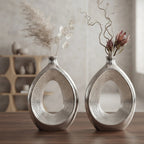 Two matching metallic silver decorative vases with an open teardrop shape, displayed on a wooden table in a softly lit interior setting with dried floral arrangements.