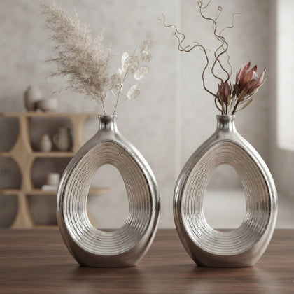 Two matching metallic silver decorative vases with an open teardrop shape, displayed on a wooden table in a softly lit interior setting with dried floral arrangements.