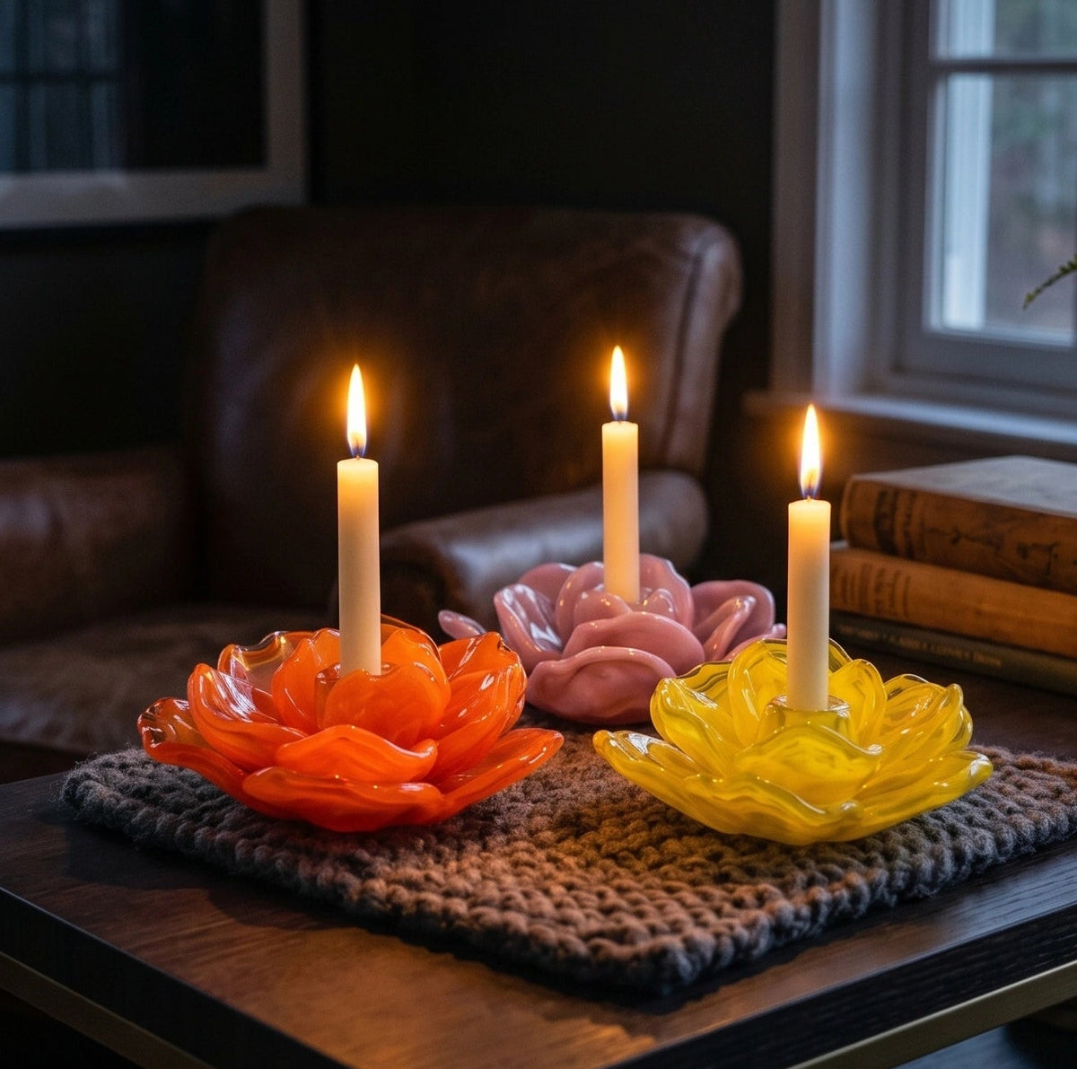Three lotus-shaped glass candle holders in orange, pink, and yellow, each holding a lit white taper candle, arranged on a knitted brown mat on a dark wooden table in a cozy indoor setting.