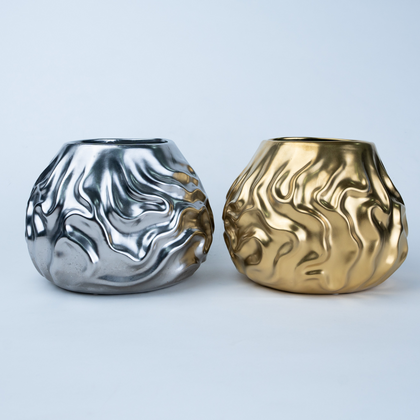 Two metallic sculptural vases with organic wavy textures, one in polished silver and one in polished gold, displayed side-by-side on a light gray background.