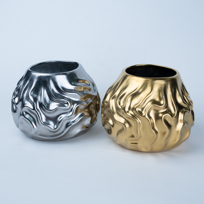 Two sculptural metallic vases with organic, wavy textured surfaces, one in polished silver and one in polished gold, set against a clean light gray background.