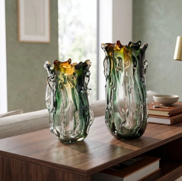 Two tall, organic-shaped glass vases with green, amber, and clear swirls, displayed on a dark wood console table in a modern living room setting.