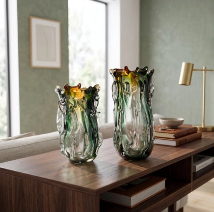 Two tall, organic-shaped glass vases with green, amber, and clear swirls, displayed on a dark wood console table in a modern living room setting.