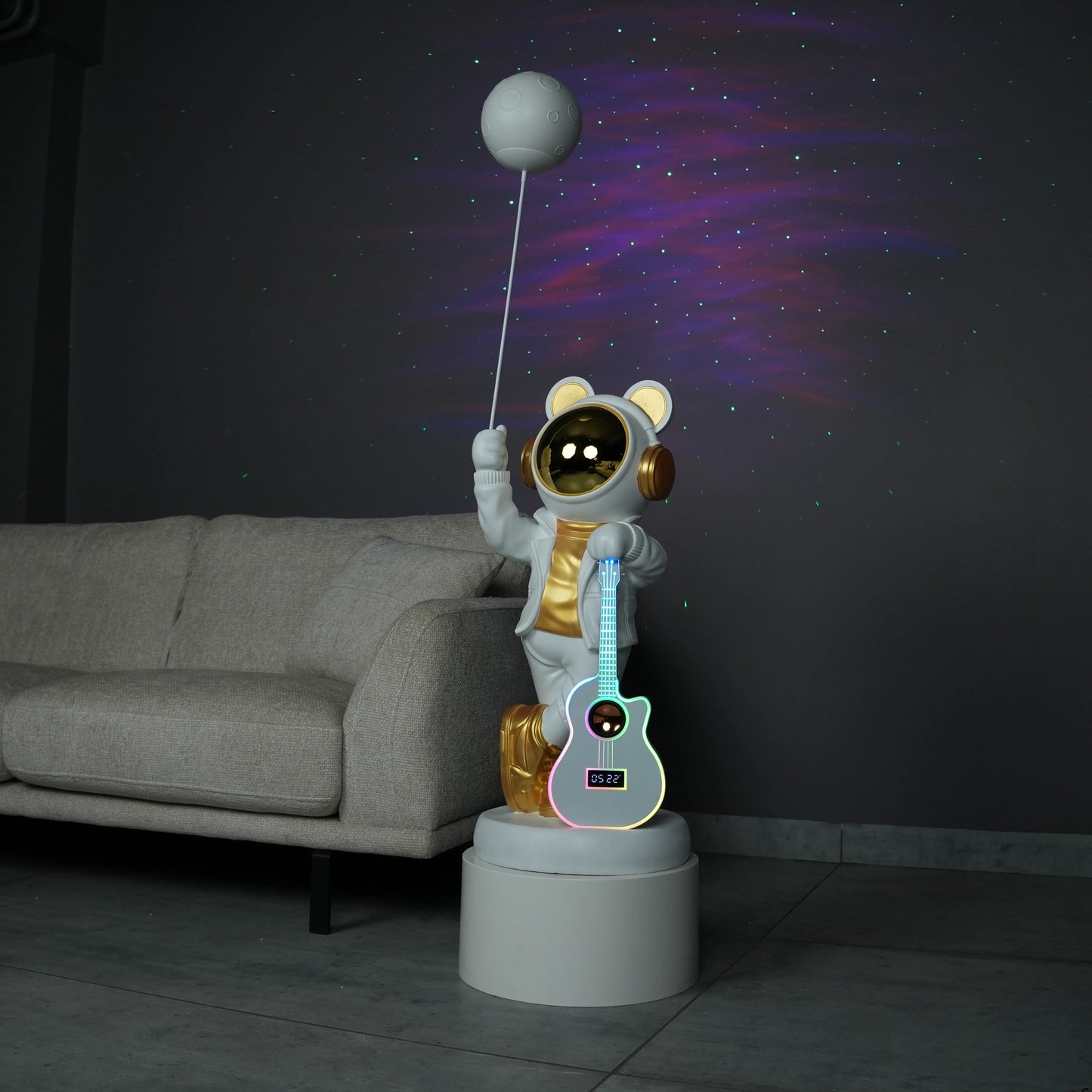 White-and-gold astronaut statue holding a moon projector balloon with a guitar-shaped Bluetooth speaker, standing on a pedestal with galaxy projection in the background.