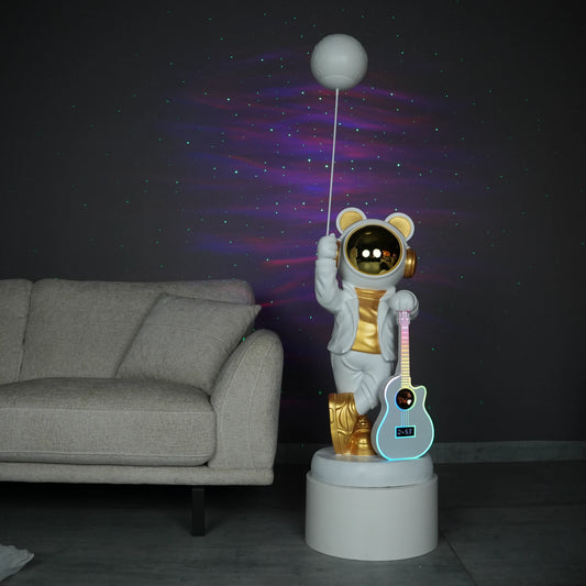 White-and-gold astronaut statue holding a moon projector balloon with a guitar-shaped Bluetooth speaker, standing on a pedestal with galaxy projection in the background.