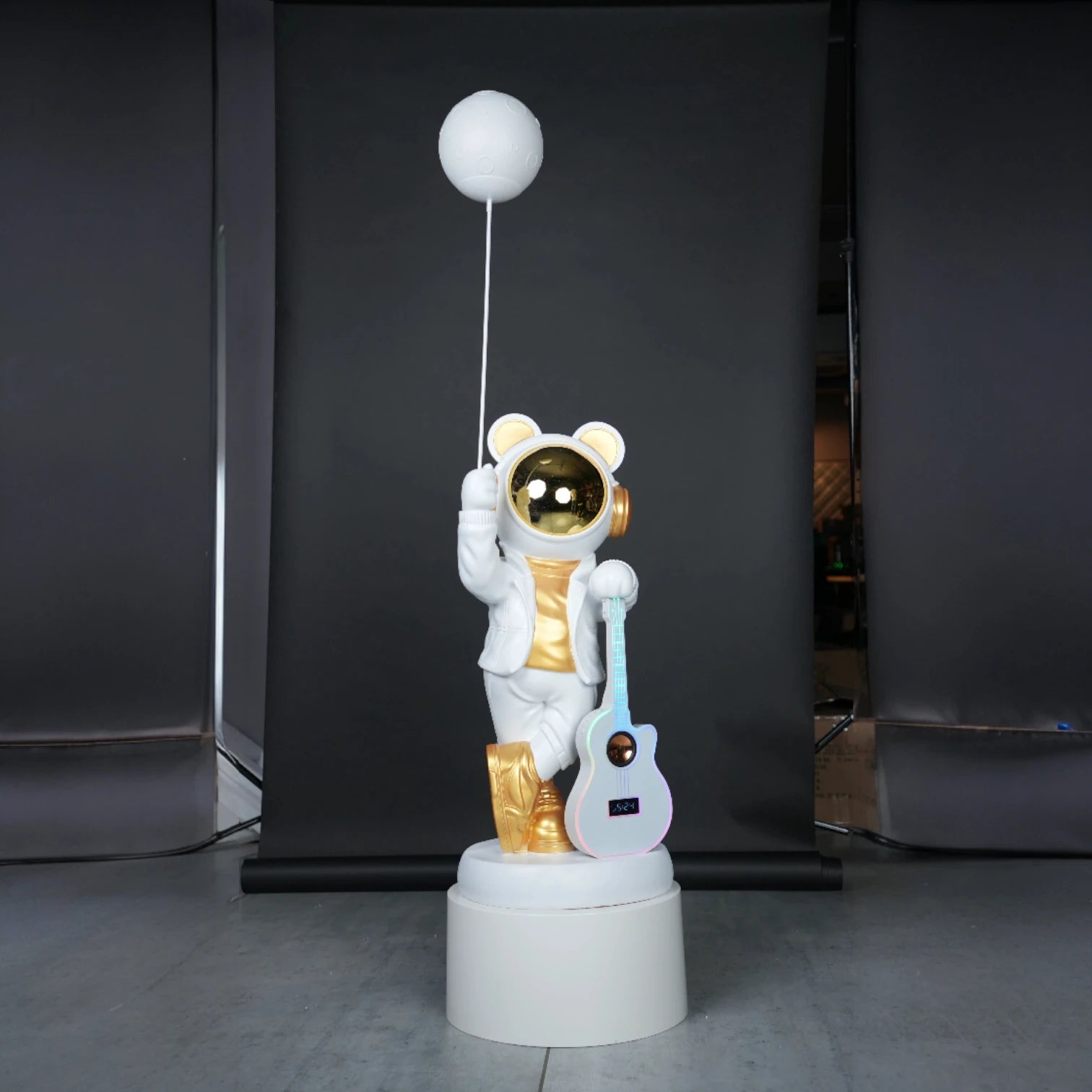 White-and-gold astronaut statue holding a moon projector balloon with a guitar-shaped Bluetooth speaker, standing on a pedestal with galaxy projection in the background.
