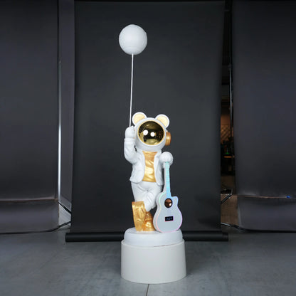 White-and-gold astronaut statue holding a moon projector balloon with a guitar-shaped Bluetooth speaker, standing on a pedestal with galaxy projection in the background.