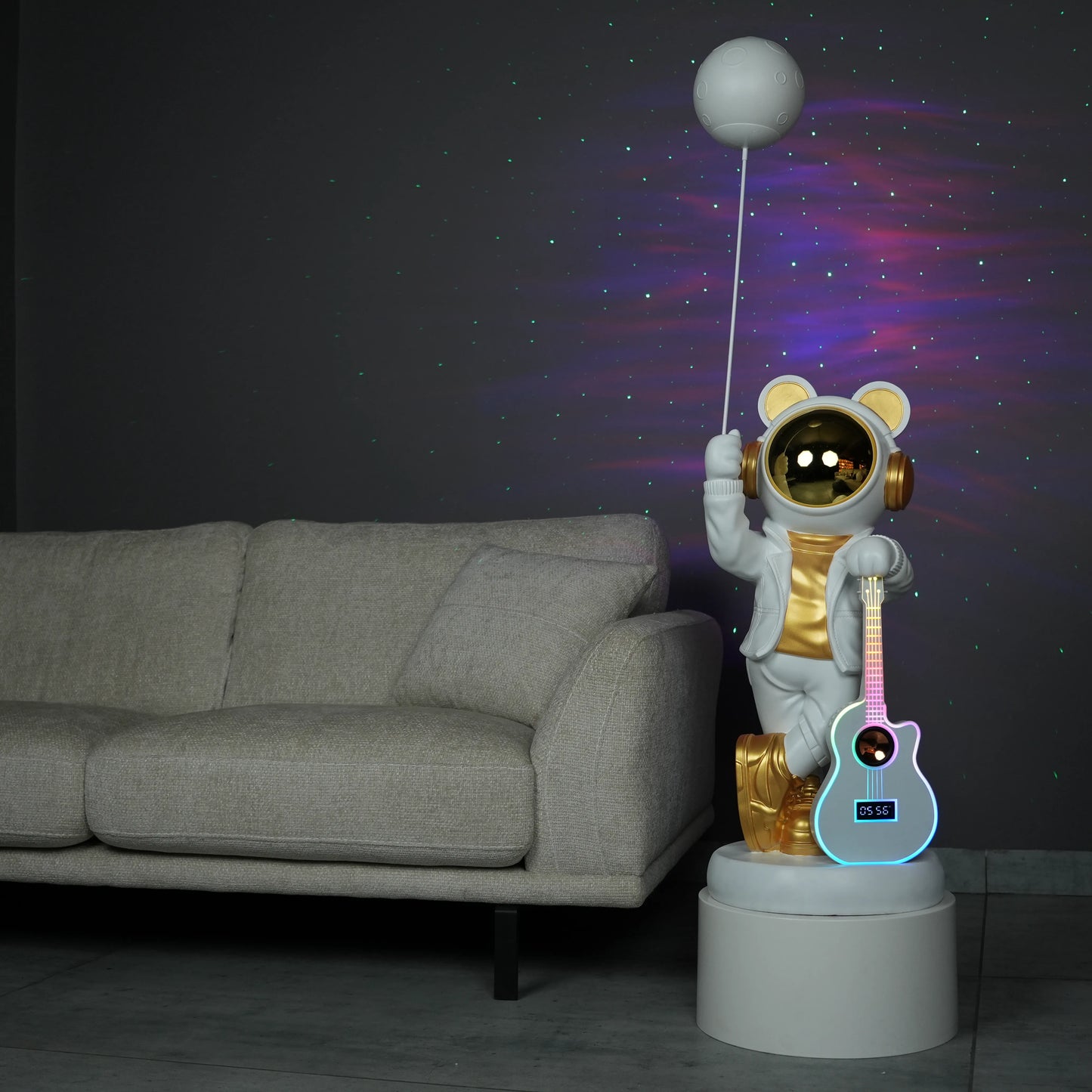 White-and-gold astronaut statue holding a moon projector balloon with a guitar-shaped Bluetooth speaker, standing on a pedestal with galaxy projection in the background.