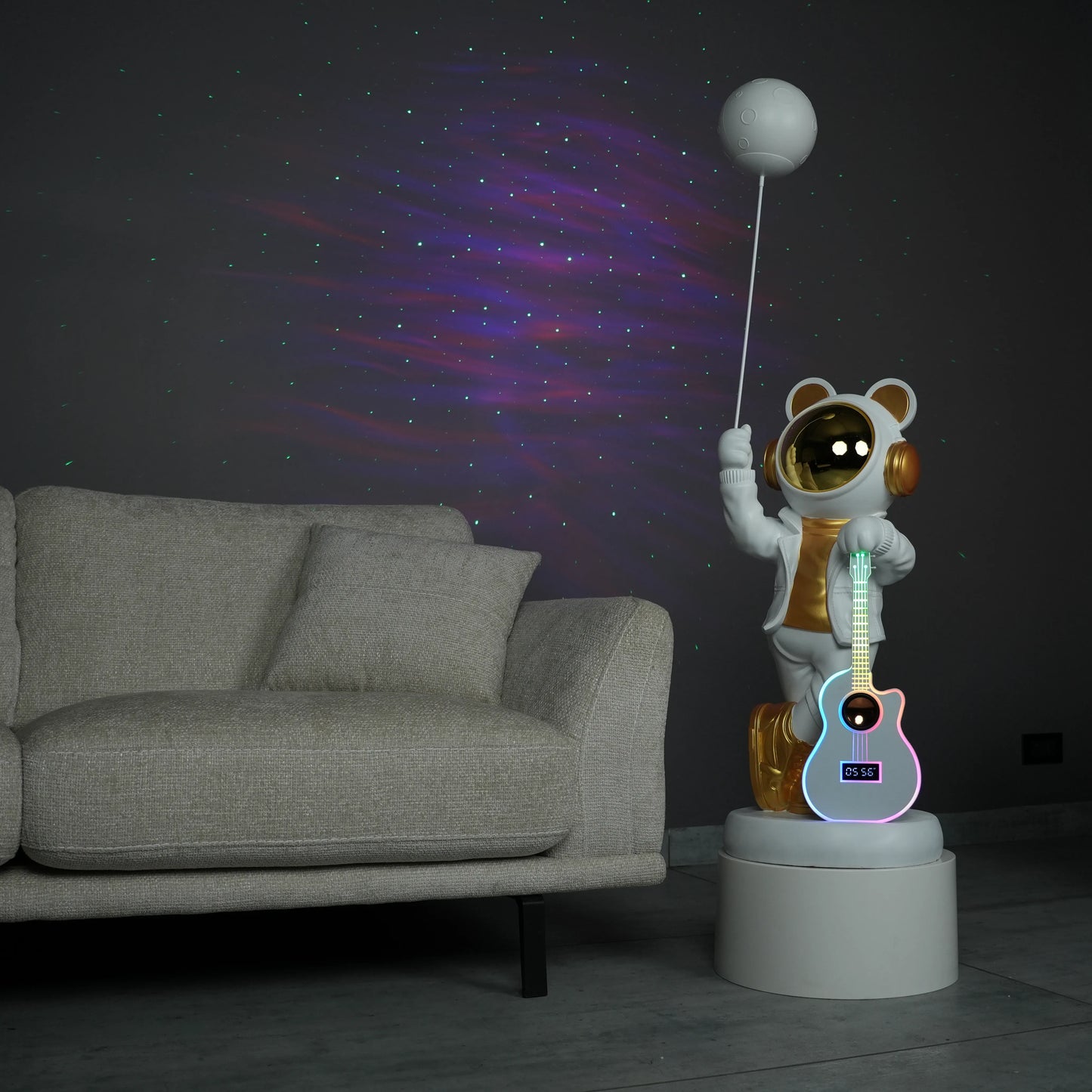White-and-gold astronaut statue holding a moon projector balloon with a guitar-shaped Bluetooth speaker, standing on a pedestal with galaxy projection in the background.